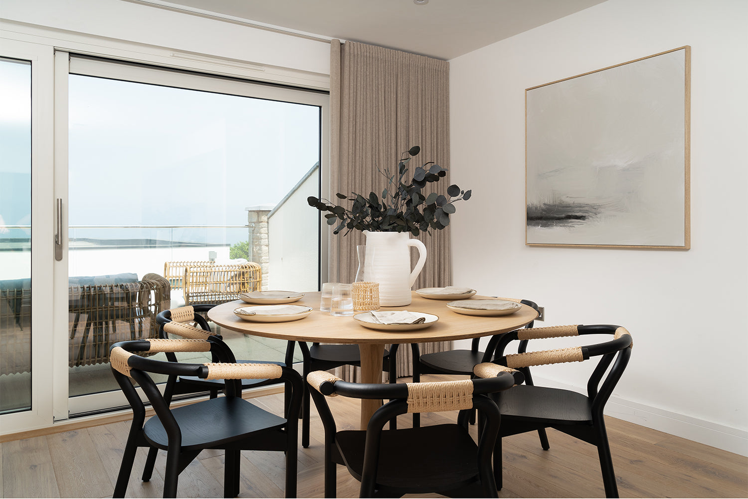 Scandi-inspired dining area at Azure showhome, Carbis Bay – featuring a round oak table, woven armchairs, abstract art and coastal views through sliding glass doors – interior styling by Iroka.