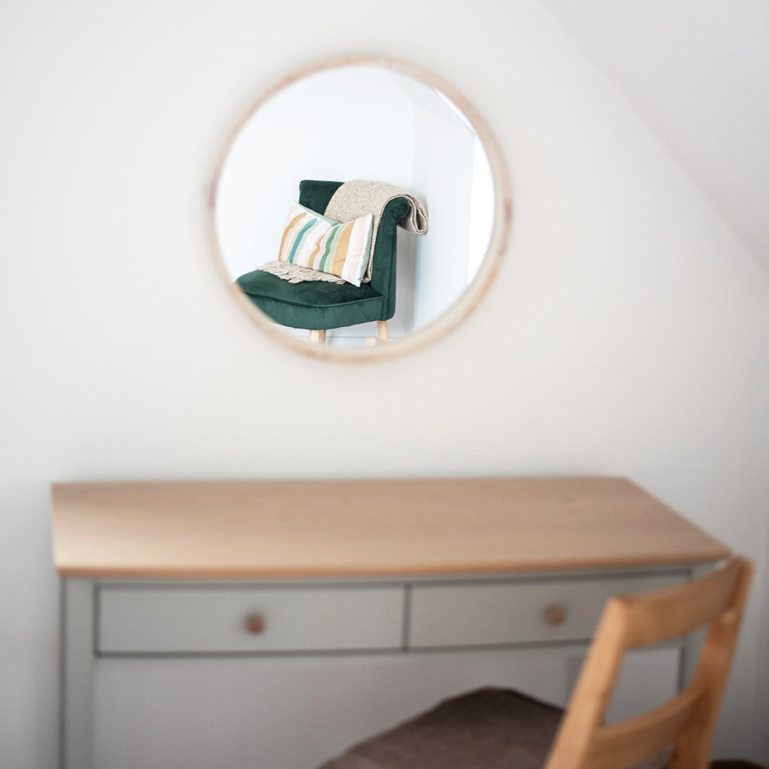 Round mirror on a wall above a wooden desk with a chair.