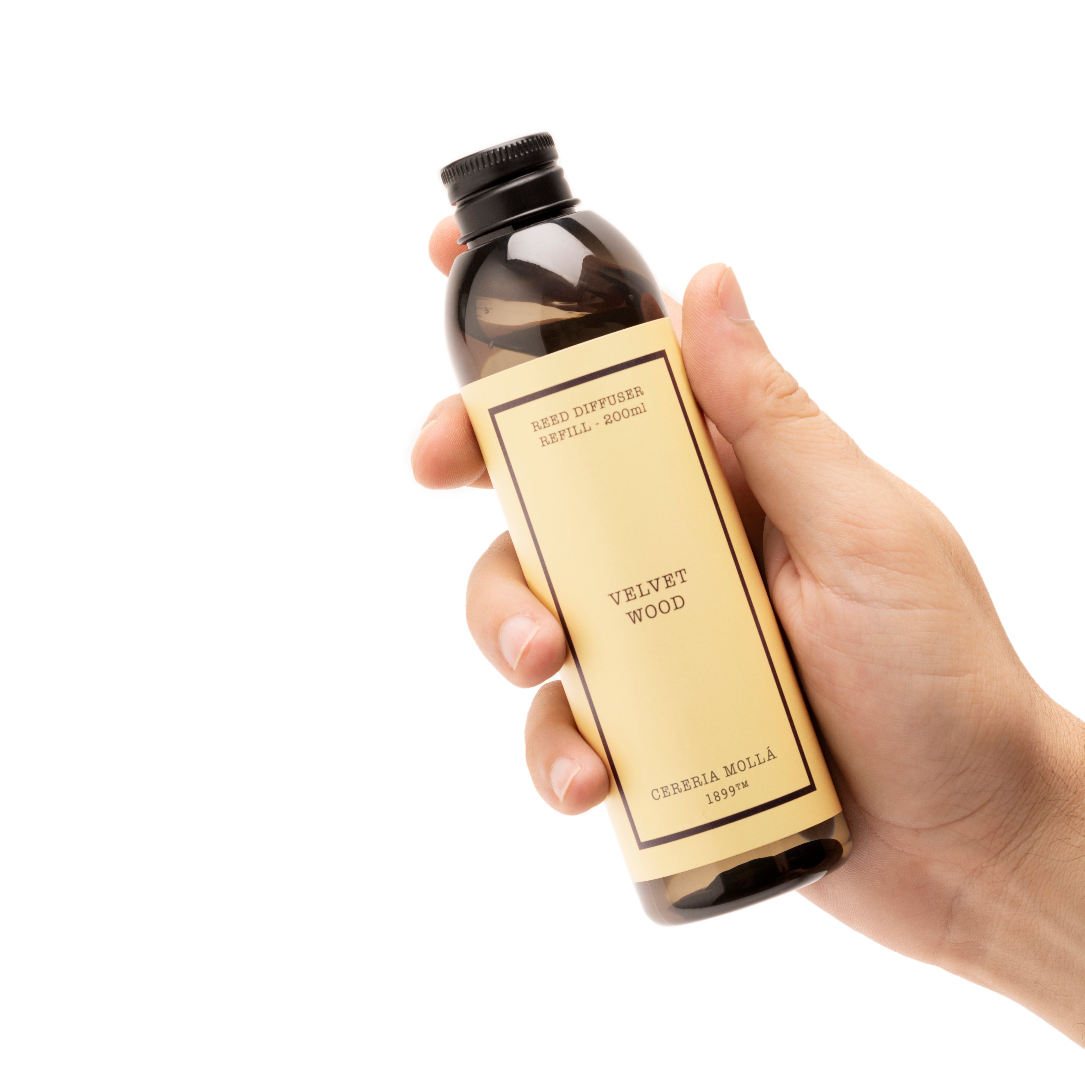 A hand holding a dark amber glass bottle with a black cap and a beige label that reads 'VELVET WOOD' and 'cereria mola' on a plain white background.