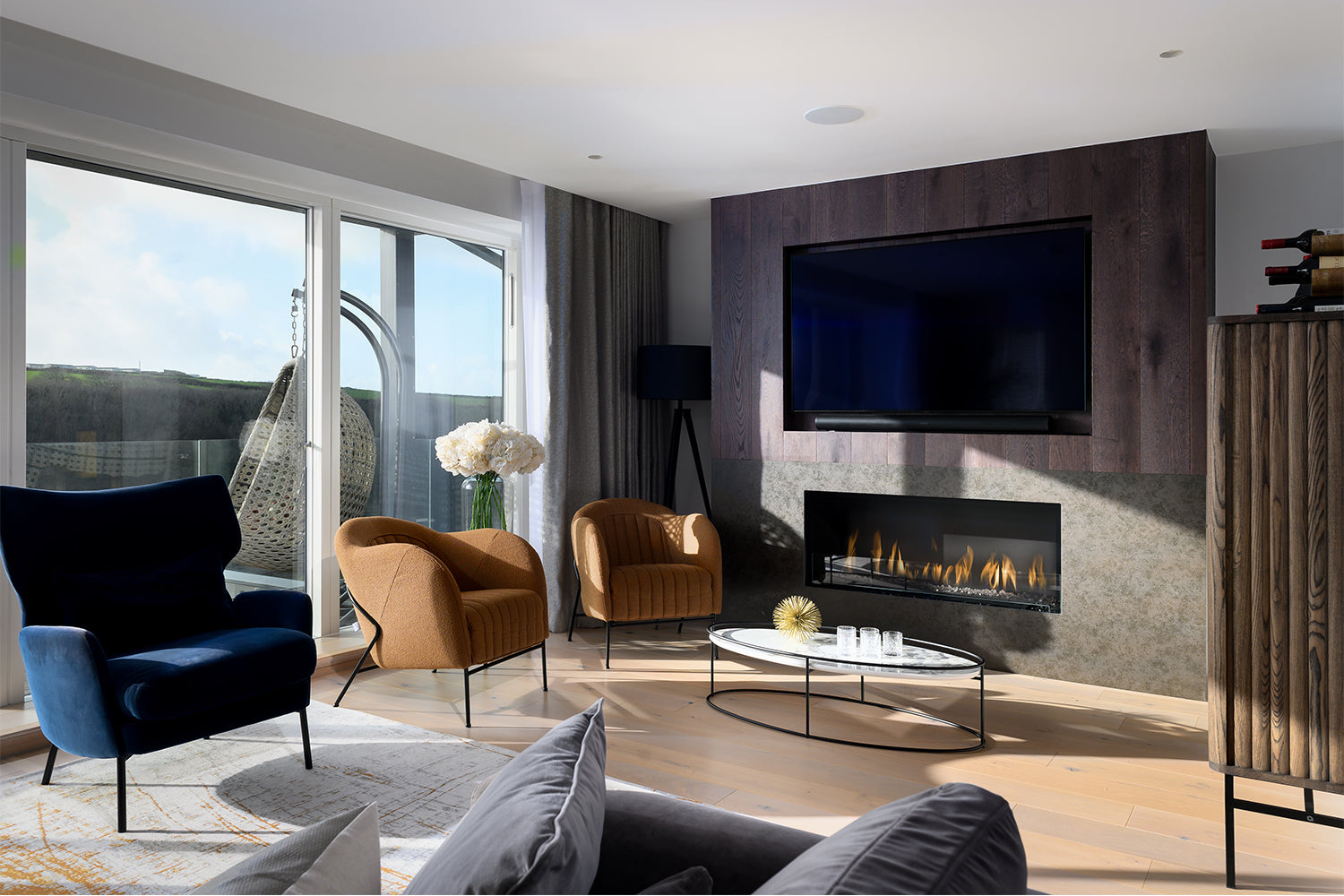 Modern coastal living room at Paradise View with panoramic sliding glass doors, contemporary velvet armchairs, built-in fireplace, and wall-mounted TV – styled by Iroka Interiors.