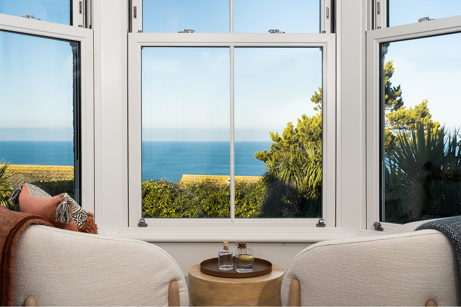 Serene window seating nook with panoramic sea views in Merryn, St Ives – plush cream armchairs, brass accent table, and coastal interiors by Iroka.