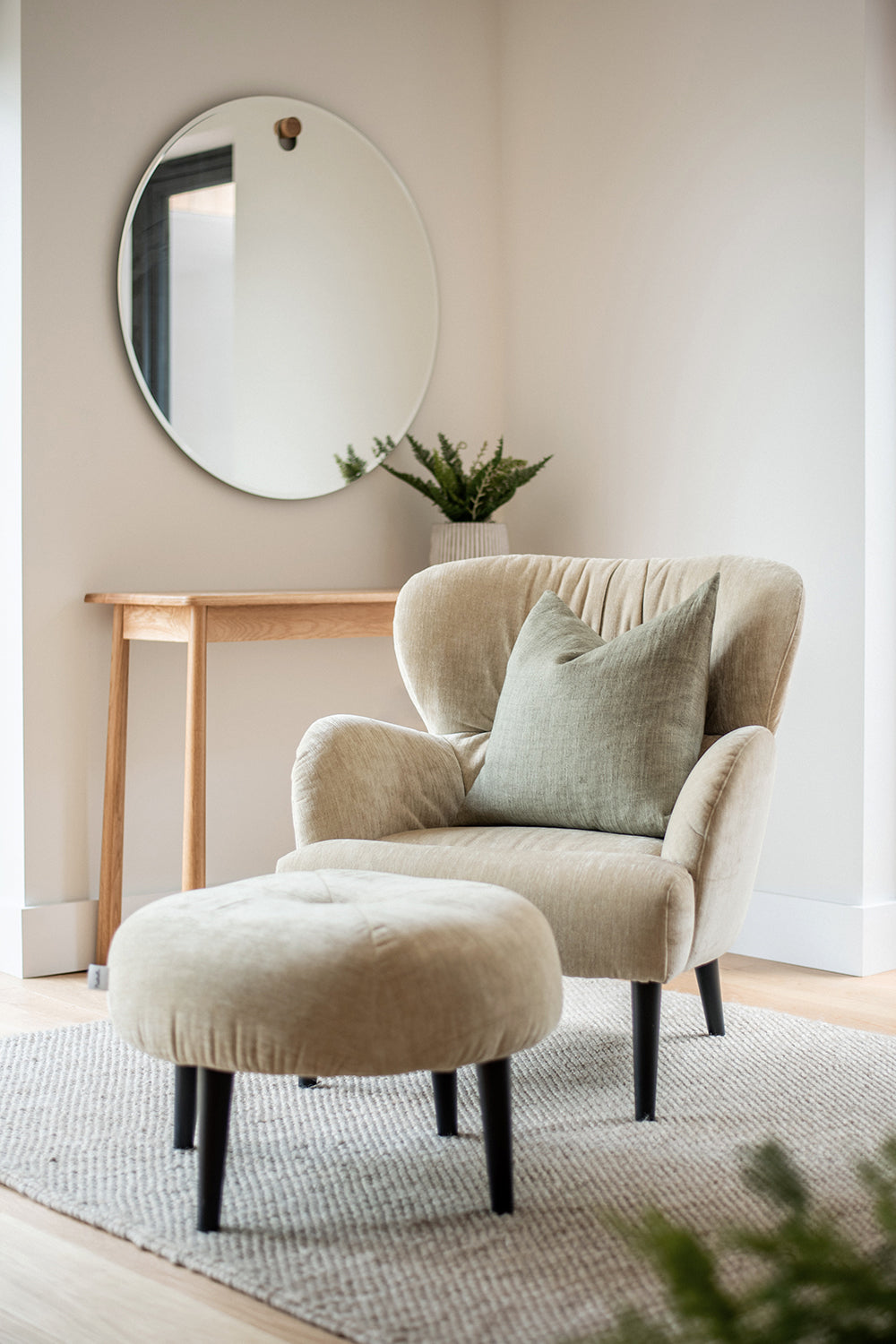 Scandinavian-style reading nook featuring a beige upholstered armchair with matching ottoman, sage green cushion, natural wood console table, round wall mirror, and neutral-toned textured rug in St Issey