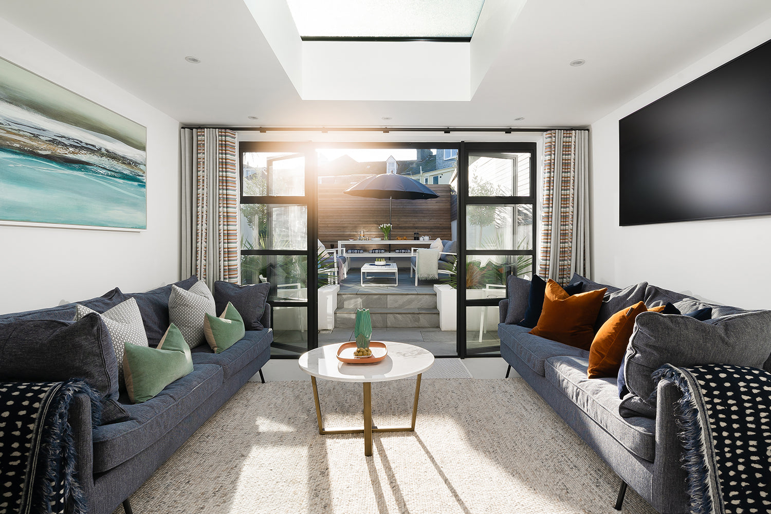 Bright, contemporary living room with twin navy sofas, colourful cushions, large skylight, and bi-fold doors opening to a sunlit balcony – designed by Iroka Interiors.