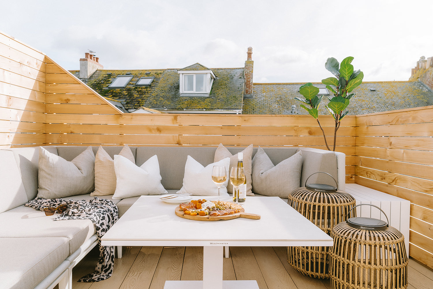 Westminster outdoor furniture on a rooftop terrace at Seaglass House, styled by Iroka Interiors with a built-in corner sofa, scatter cushions, rattan lanterns, and a white dining table set for wine and charcuterie.