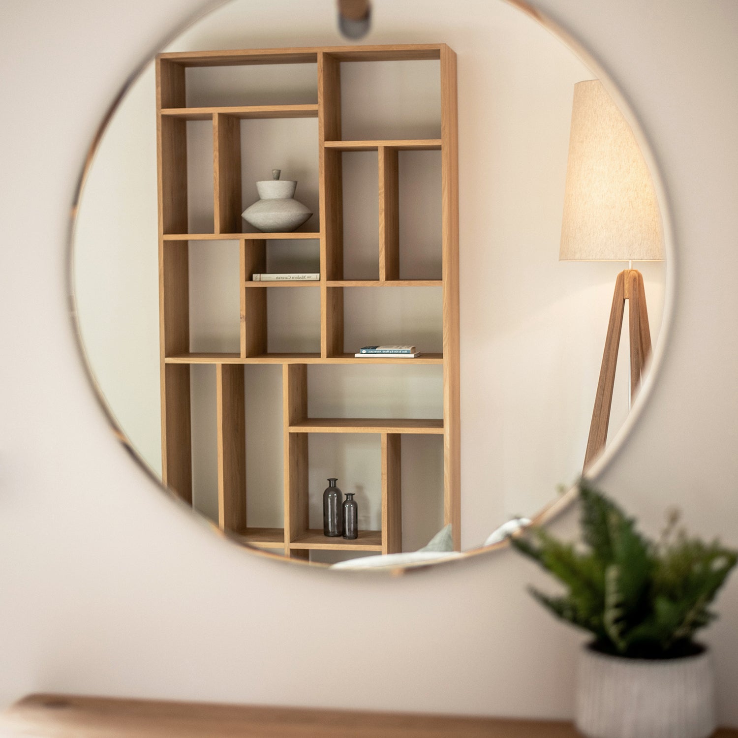Modern wooden bookshelf with geometric open shelving reflected in a round wall mirror, styled with minimalist decor including ceramic vases and books; accompanied by a tripod floor lamp in a neutral-toned interior.