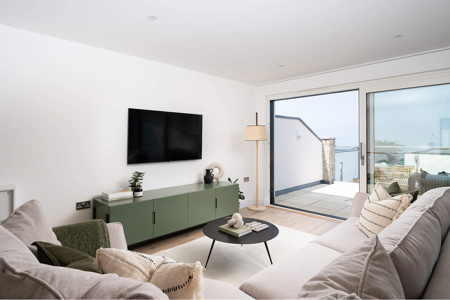 Modern coastal living room at Azure showhome, Carbis Bay – features a green fluted sideboard, wall-mounted TV, boucle sofas and sea views through sliding doors – styled by Iroka Interiors.
