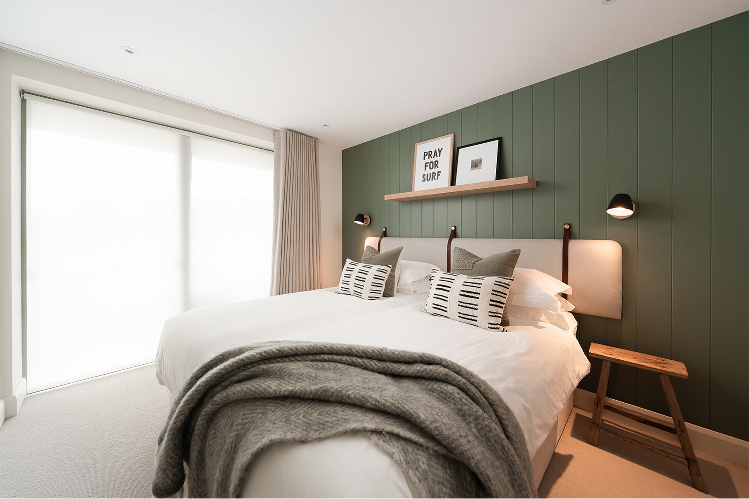 Modern coastal bedroom in Polzeath with sage green feature wall, layered neutral bedding, floating shelf styling and soft natural light – interiors by Iroka.