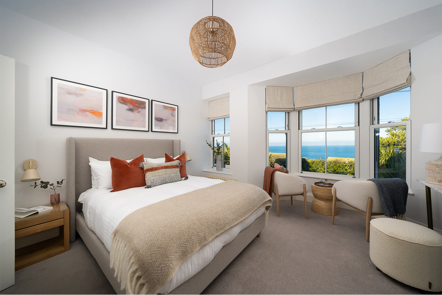 Bright coastal bedroom interior with neutral palette, terracotta accents, modern wall art, bay windows overlooking the ocean, and contemporary armchairs – styled by Iroka Interiors, St Ives.