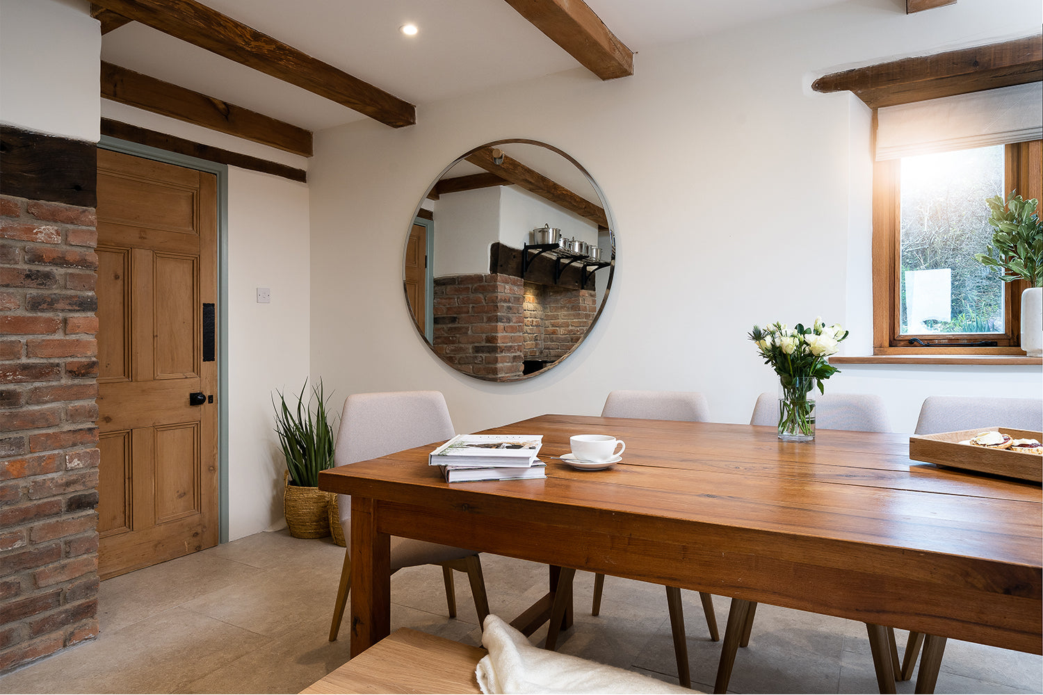 Rustic dining room with exposed beams, natural wood table, stone wall, and round feature mirror – interior design by Iroka Interiors in Cornwall.