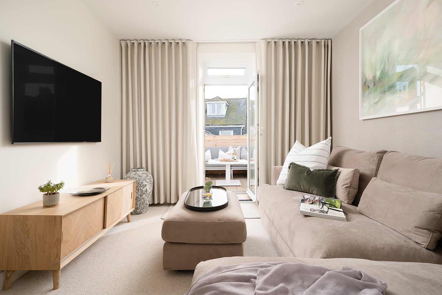 Interior view of Seaglass House styled by Iroka Interiors, featuring a neutral-toned living room with a soft taupe sofa, layered cushions, natural wood sideboard, and full-height curtains framing a view of the rooftop terrace with Westminster outdoor furniture.