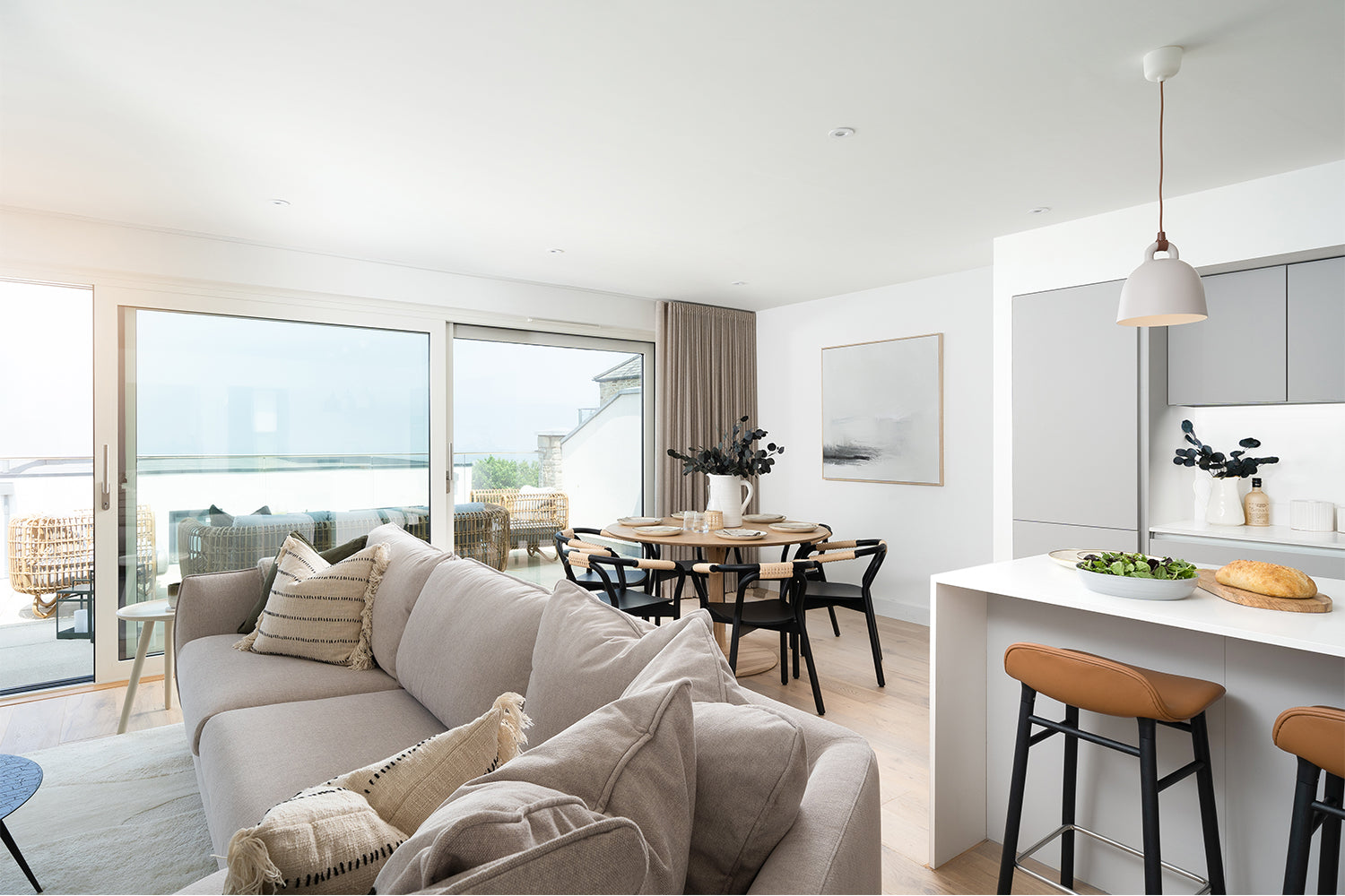 Open-plan living space at Azure showhome, Carbis Bay – features a soft neutral corner sofa, minimalist kitchen island with leather bar stools, and a round dining table styled with contemporary decor and sea views through sliding glass doors – designed by Iroka Interiors.