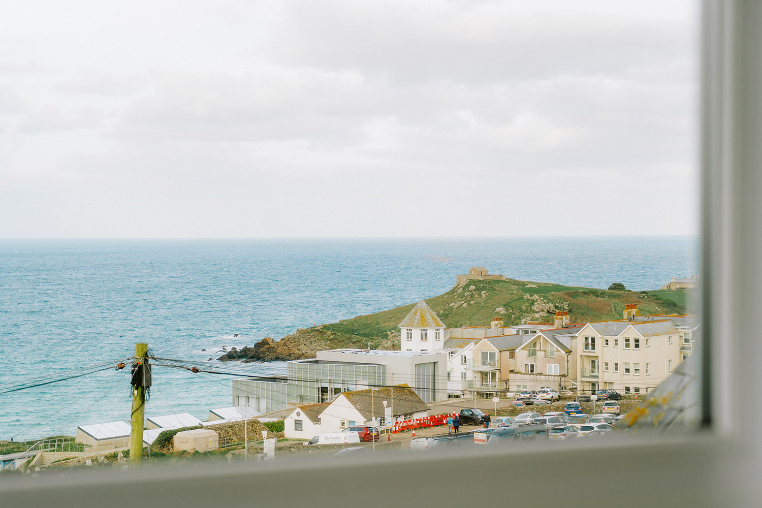 Seaglass House coastal view through a window, overlooking the turquoise sea and the Tate St Ives gallery with the iconic headland of The Island in the background, captured during an Iroka Interiors design project.