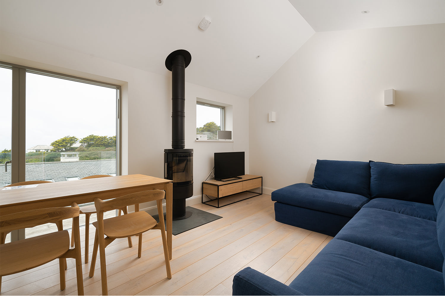 Modern Cornish living room with navy blue corner sofa, Scandinavian-style oak dining table, wood-burning stove and minimalist decor - interior by Iroka.
