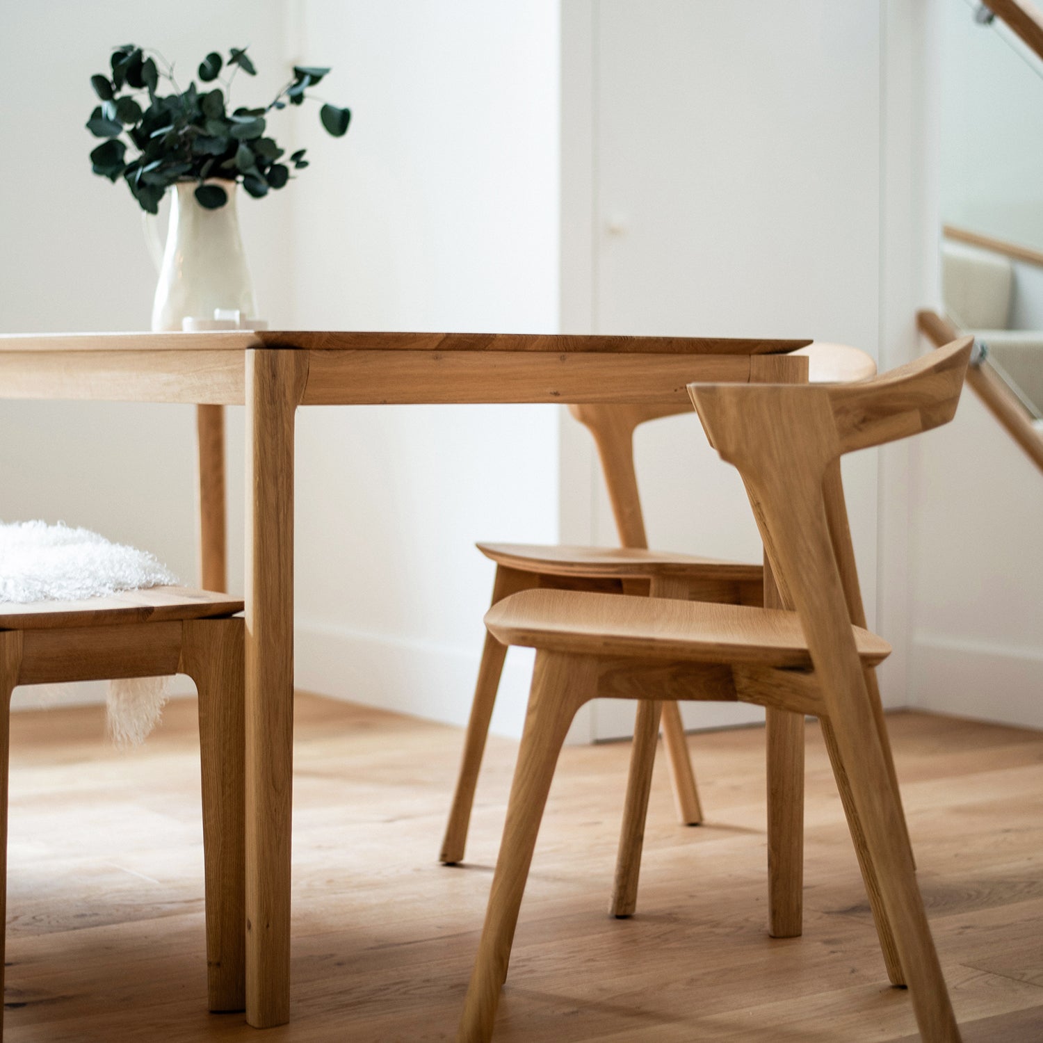 Close-up of a minimalist oak dining table with sculpted wooden chairs on a light wood floor, styled with a white ceramic vase filled with eucalyptus branches in a bright modern interior.