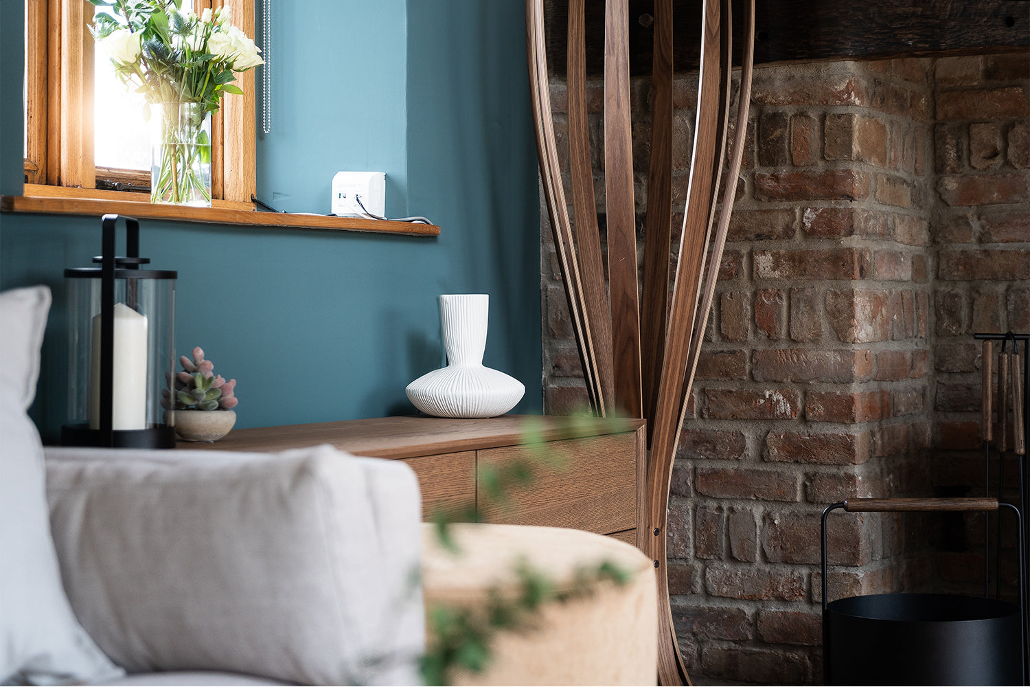 Cosy corner of a rustic living room with exposed brick fireplace, teal feature wall, and natural wood sideboard styled by Iroka Interiors, Cornwall.
