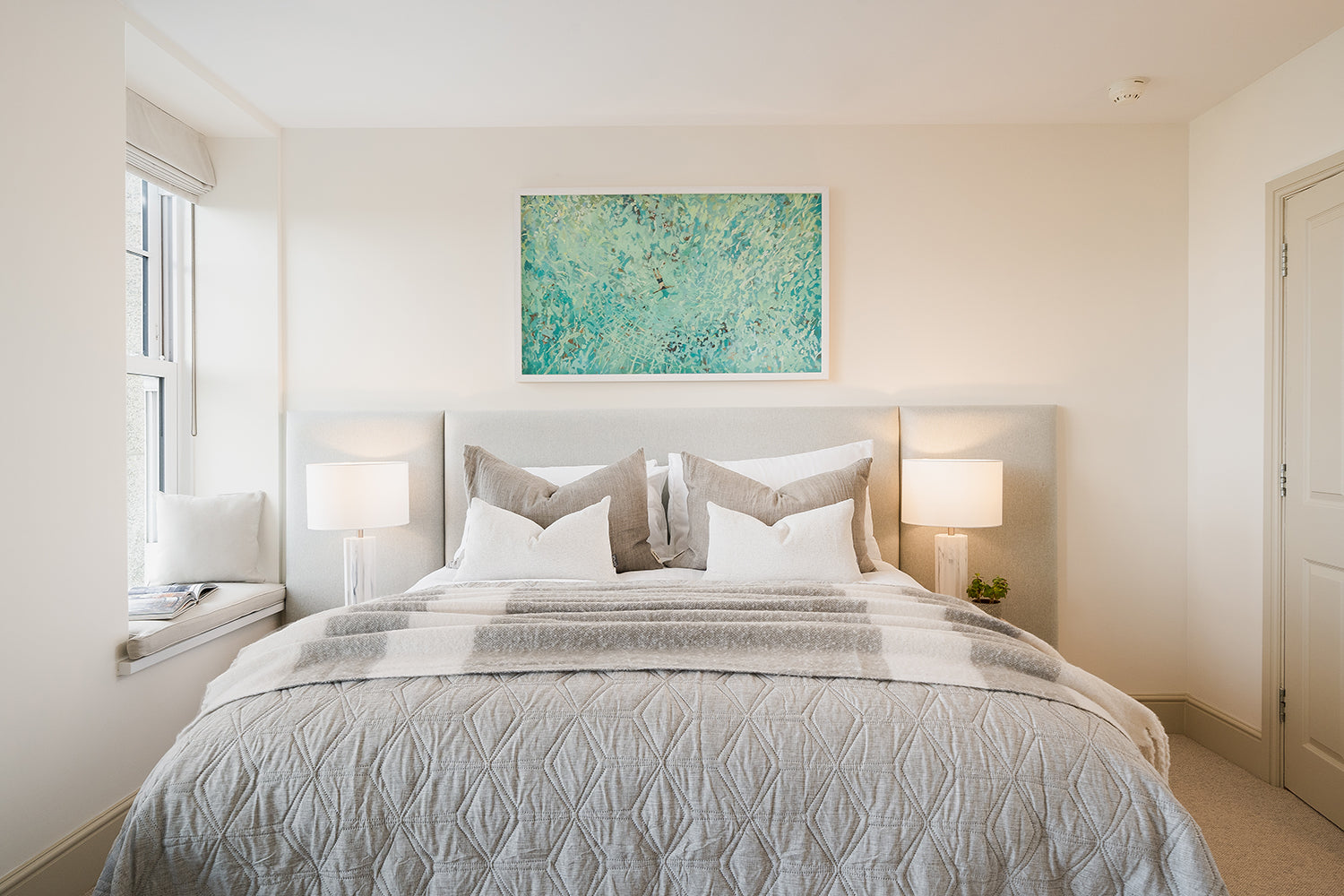 Bright coastal bedroom interior designed by Iroka Interiors, featuring a soft grey upholstered bed, layered linen bedding, matching bedside lamps, and a vibrant sea-inspired artwork above the headboard.
