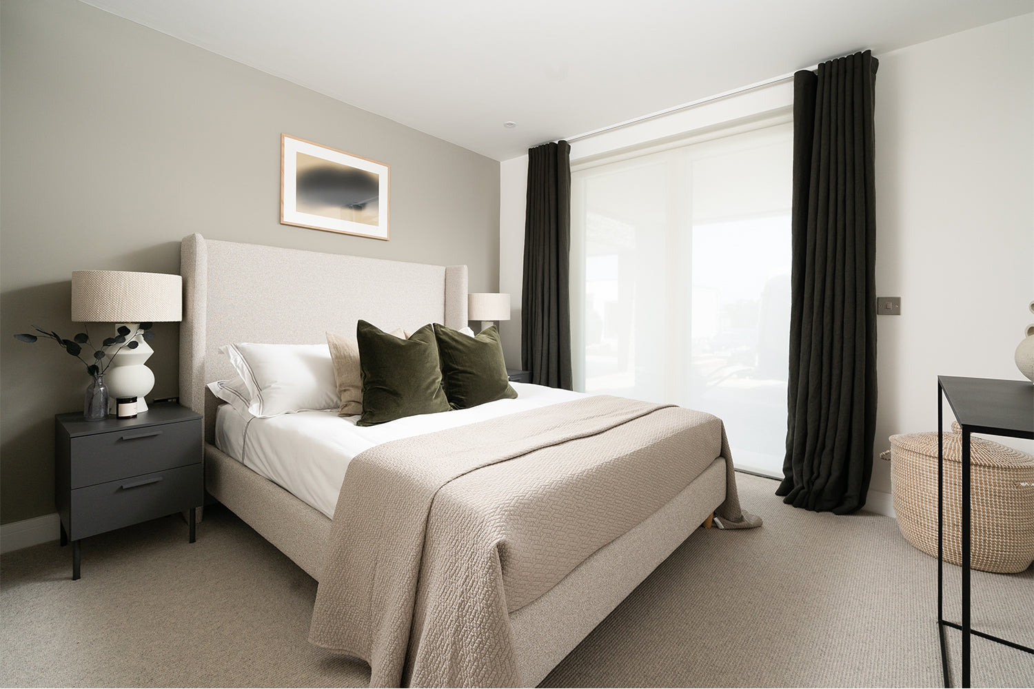 Calming modern bedroom at Azure showhome, Carbis Bay – features a soft upholstered bed, olive green and neutral cushions, sleek black bedside tables, and floor-to-ceiling curtains framing sliding glass doors – interior design by Iroka Interiors.