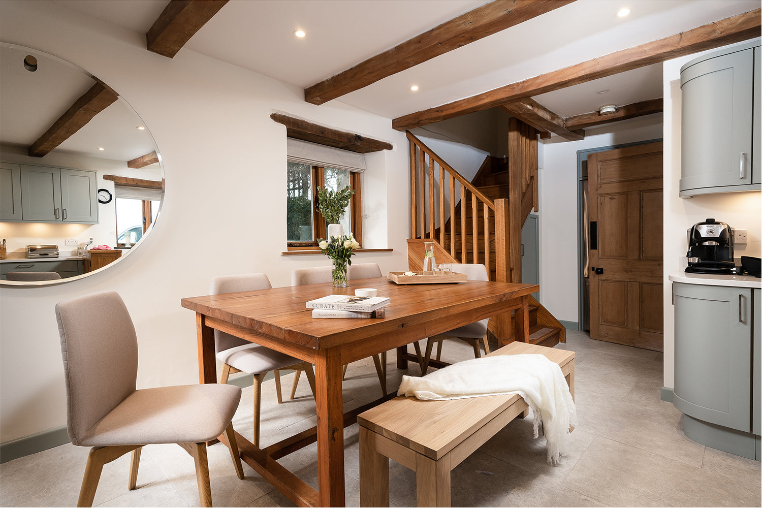 Warm rustic dining room with exposed wood beams, solid oak table, soft upholstered chairs, and sage green cabinetry – styled by Iroka Interiors in Port Isaac, Cornwall.