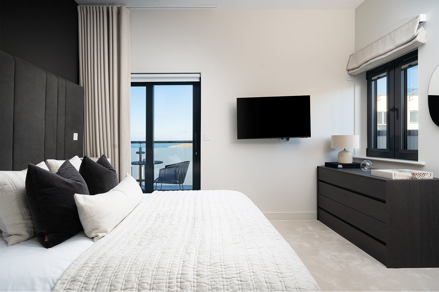 Modern coastal bedroom at Pentire Penthouse, Newquay – featuring monochrome styling, bespoke blackout headboard wall, and panoramic sea views through sliding glass doors, designed by Iroka Interiors.