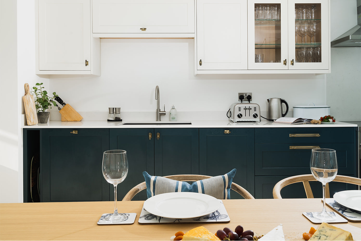 Contemporary kitchen with white upper cabinets, deep navy base units, open wood shelving, and a modern dining table set for two – styled by Iroka Interiors.