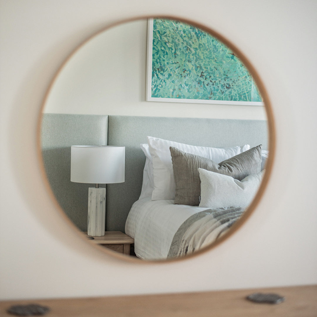 Refined bedroom reflected in round wall mirror, featuring soft sage green upholstered headboard, neutral layered pillows, marble bedside lamp, and abstract coastal artwork – serene modern interior design inspiration