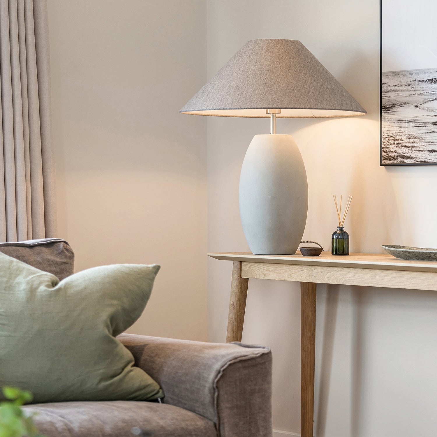 Neutral-toned living room corner with modern ceramic table lamp on light oak console table, soft grey sofa with green cushion, wall art, and minimalist decor – cozy and contemporary home styling inspiration.