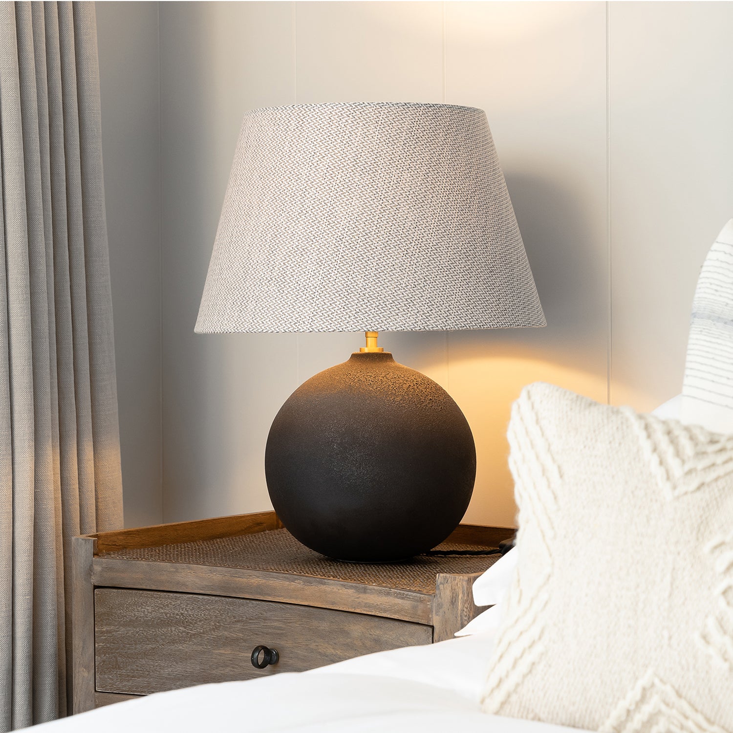 Contemporary bedside setup featuring a textured black ceramic table lamp with a woven fabric lampshade, placed on a rustic wooden nightstand beside a cream knit cushion and neutral curtains.