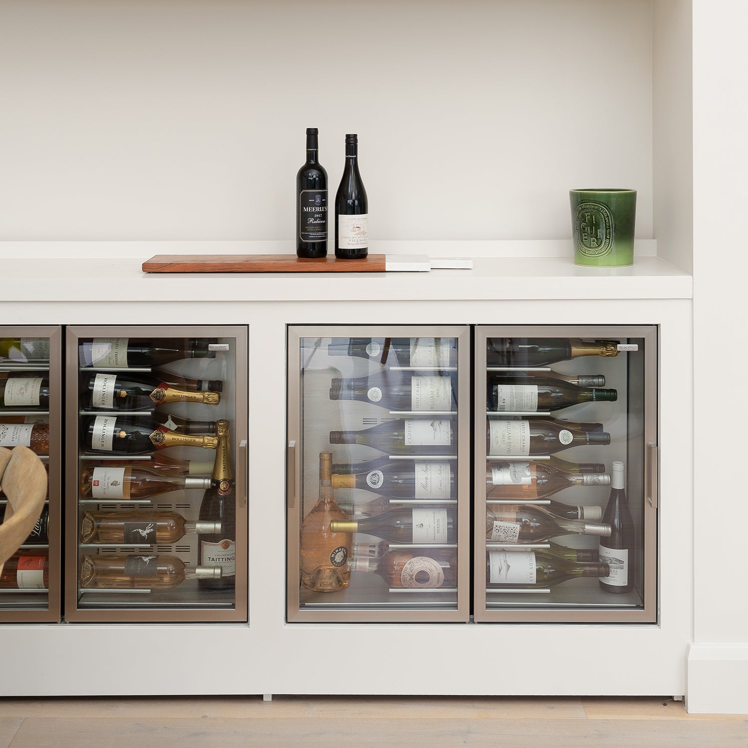 Modern built-in wine cooler with triple glass doors housing assorted wine and champagne bottles, topped with minimalist decor including two wine bottles and a green Diptyque candle.