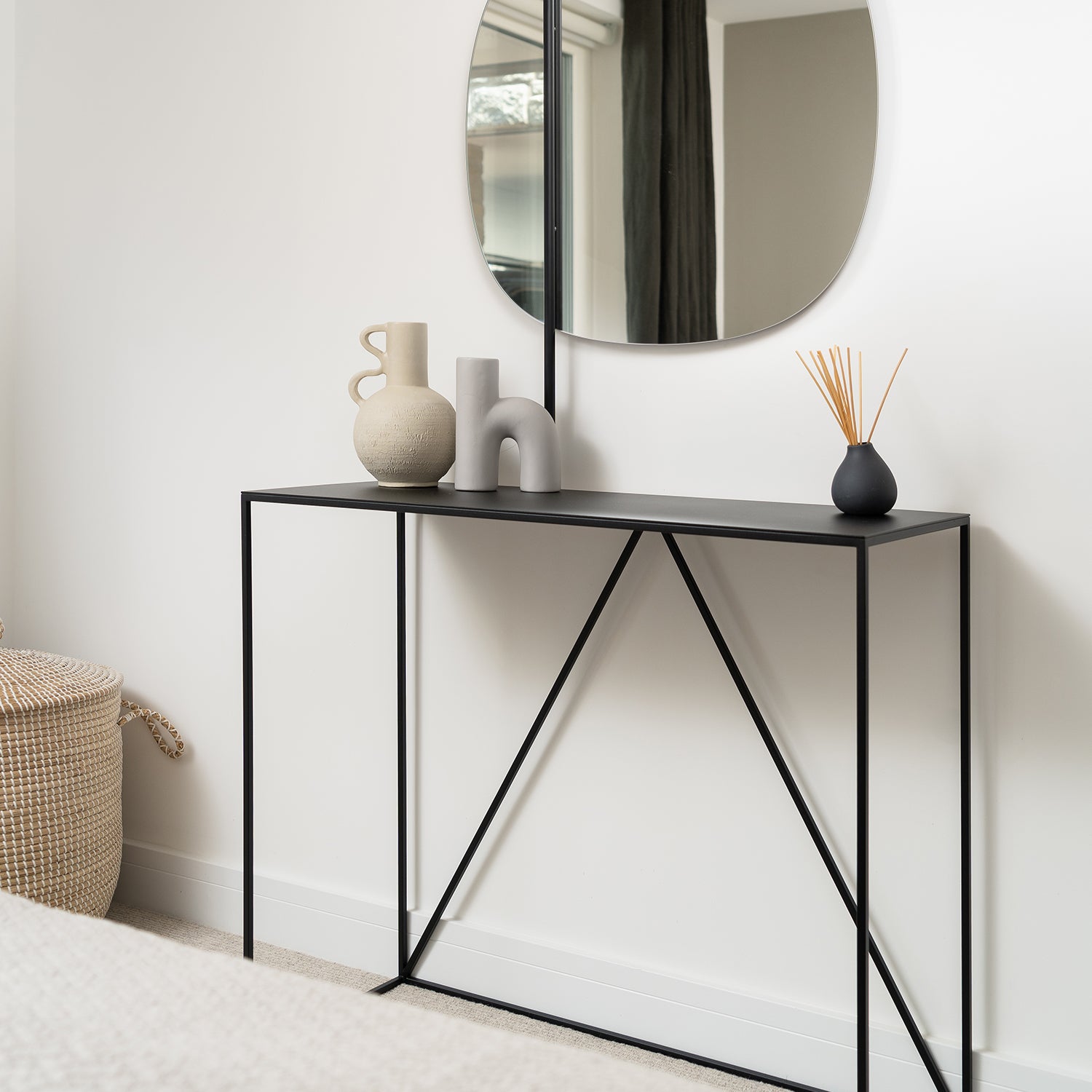Minimalist black metal console table styled with modern ceramic vases, a black reed diffuser, and an organic round mirror above; set against a white wall with neutral decor accents and a woven basket in the corner.