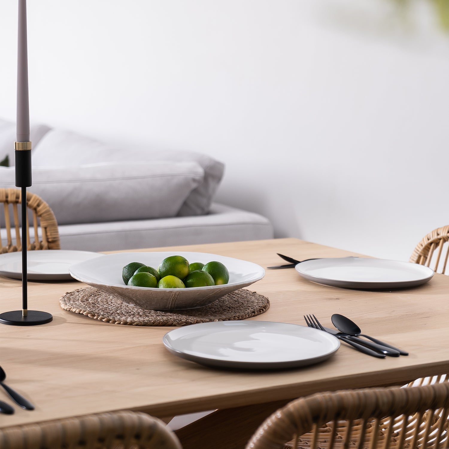 Modern natural wood dining table set with white ceramic plates, matte black cutlery, and a woven placemat featuring a decorative bowl of fresh green limes; rattan dining chairs and a neutral-toned sofa in the background.