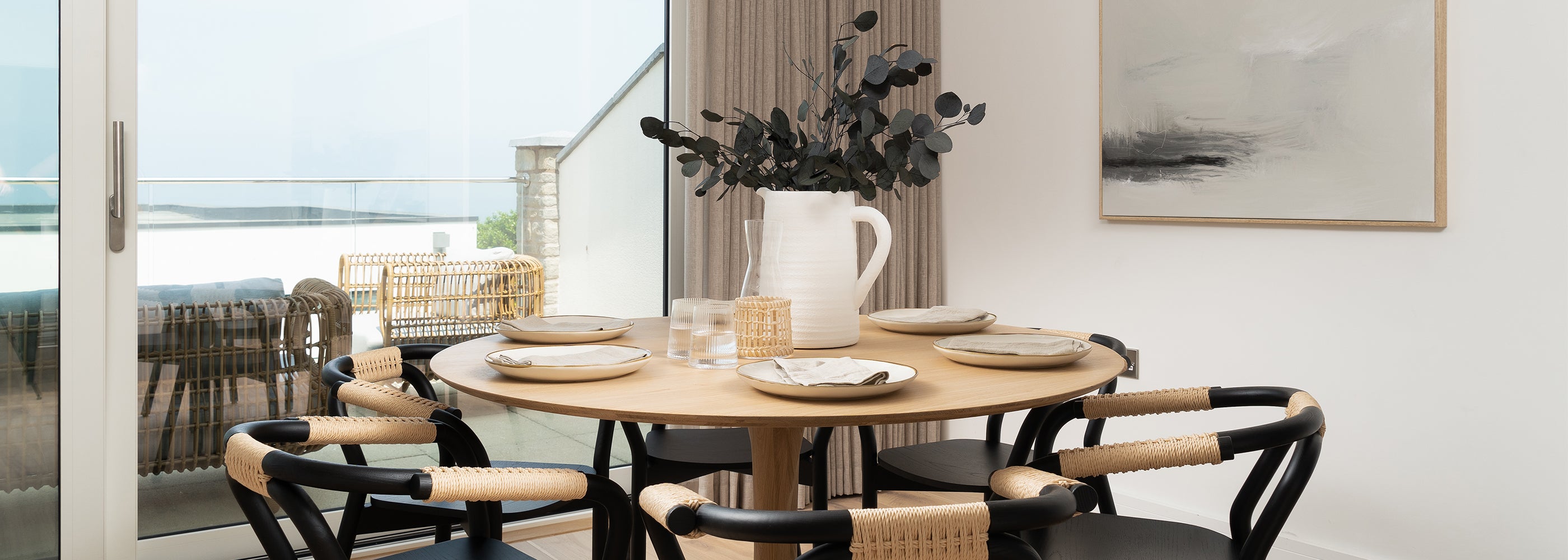 Scandinavian-style dining space by Iroka Interiors in Hayle, Cornwall featuring a round natural oak table, modern black and rattan chairs, and minimalist decor with coastal views through glass sliding doors – perfect for relaxed contemporary living.