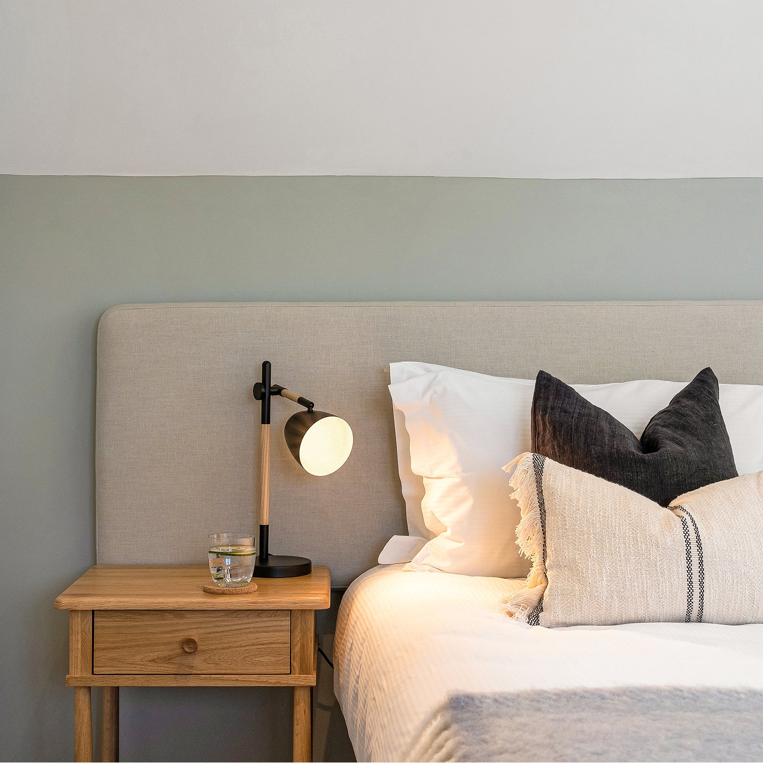 Contemporary bedroom scene with a light wood bedside table, black and wood reading lamp, beige upholstered headboard, layered neutral cushions, and two-tone painted wall in soft grey and white.