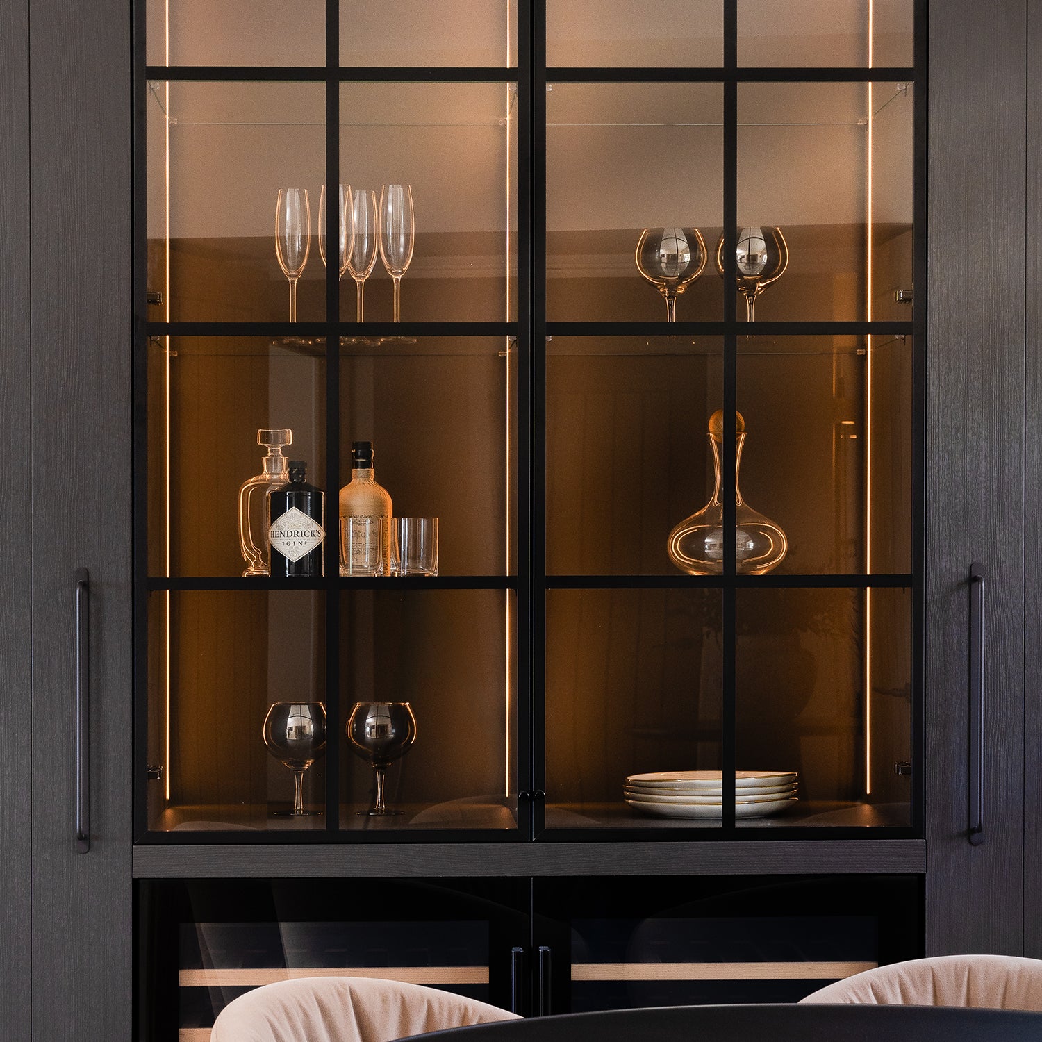 Elegant built-in kitchen bar cabinet with dark wood finish, ambient LED lighting, glass shelving, and a curated display of glassware and spirits – luxury kitchen design by Iroka Interiors in Cornwall.