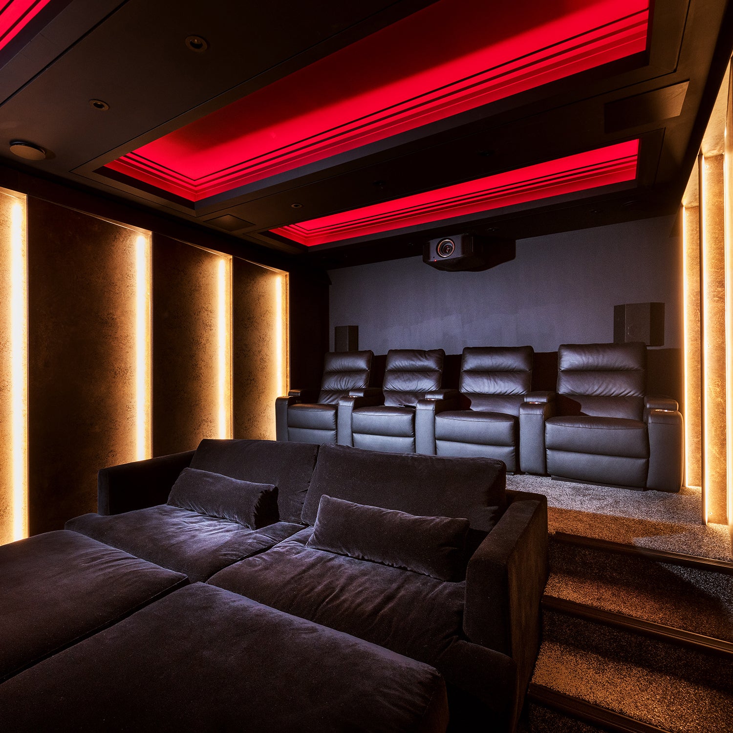 Luxury home cinema room featuring tiered seating with plush velvet recliners, dramatic red ambient ceiling lighting, and vertical LED wall panels – designed and installed by Iroka Interiors in Cornwall.