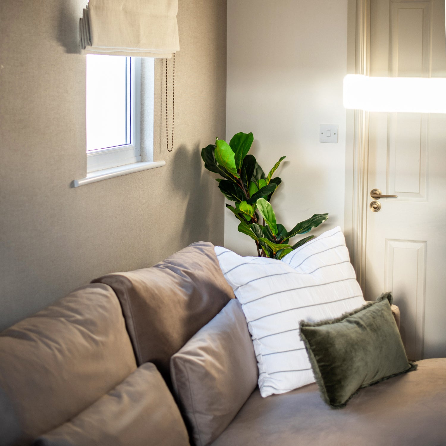 Cozy corner of a neutral-toned living space at Seaglass House, featuring a soft taupe sofa with layered cushions, a green velvet pillow, and natural light streaming through sheer curtains near a potted plant.