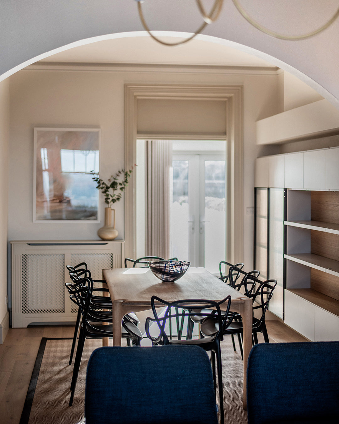Contemporary dining room at Seaglass House in St Ives, Cornwall, designed by Iroka Interiors, featuring a natural oak table with sculptural black dining chairs, arched ceiling, built-in shelving, and soft neutral tones with coastal daylight streaming through French doors.