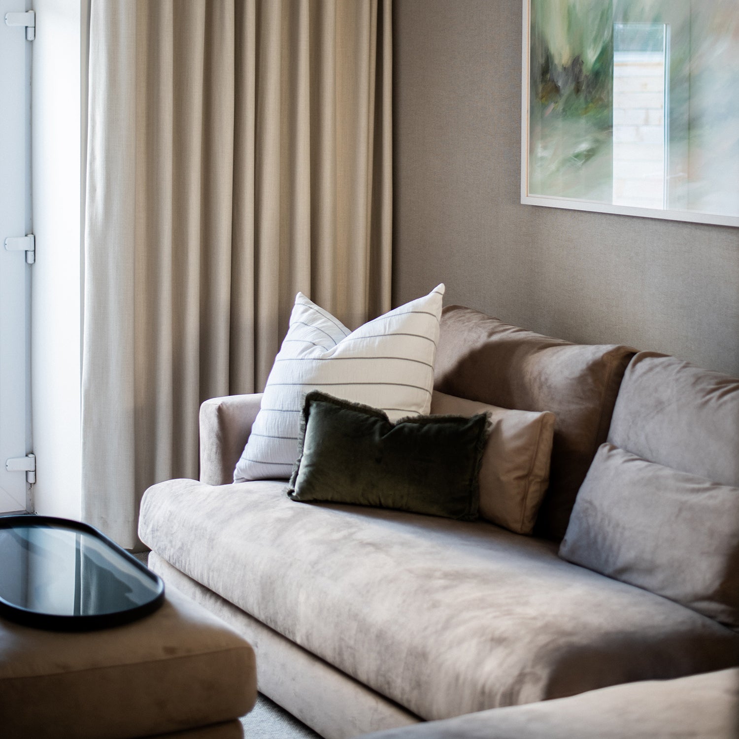 Elegant living room interior at Clodgy View in St Ives, Cornwall, featuring a plush taupe sofa with textured scatter cushions, full-height neutral curtains, and abstract wall art, designed by Iroka Interiors.