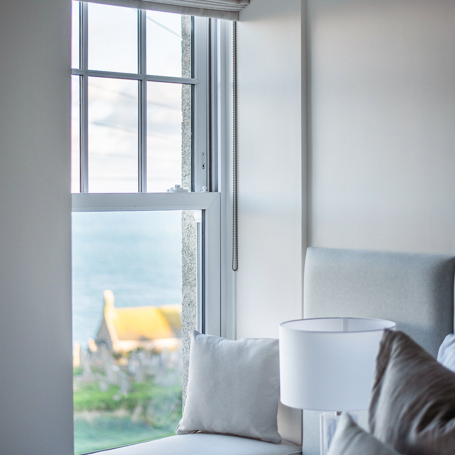 Bright and serene bedroom corner at Clodgy View in St Ives, Cornwall, featuring soft grey upholstered furnishings and a sash window overlooking the sea and countryside, styled by Iroka Interiors.
