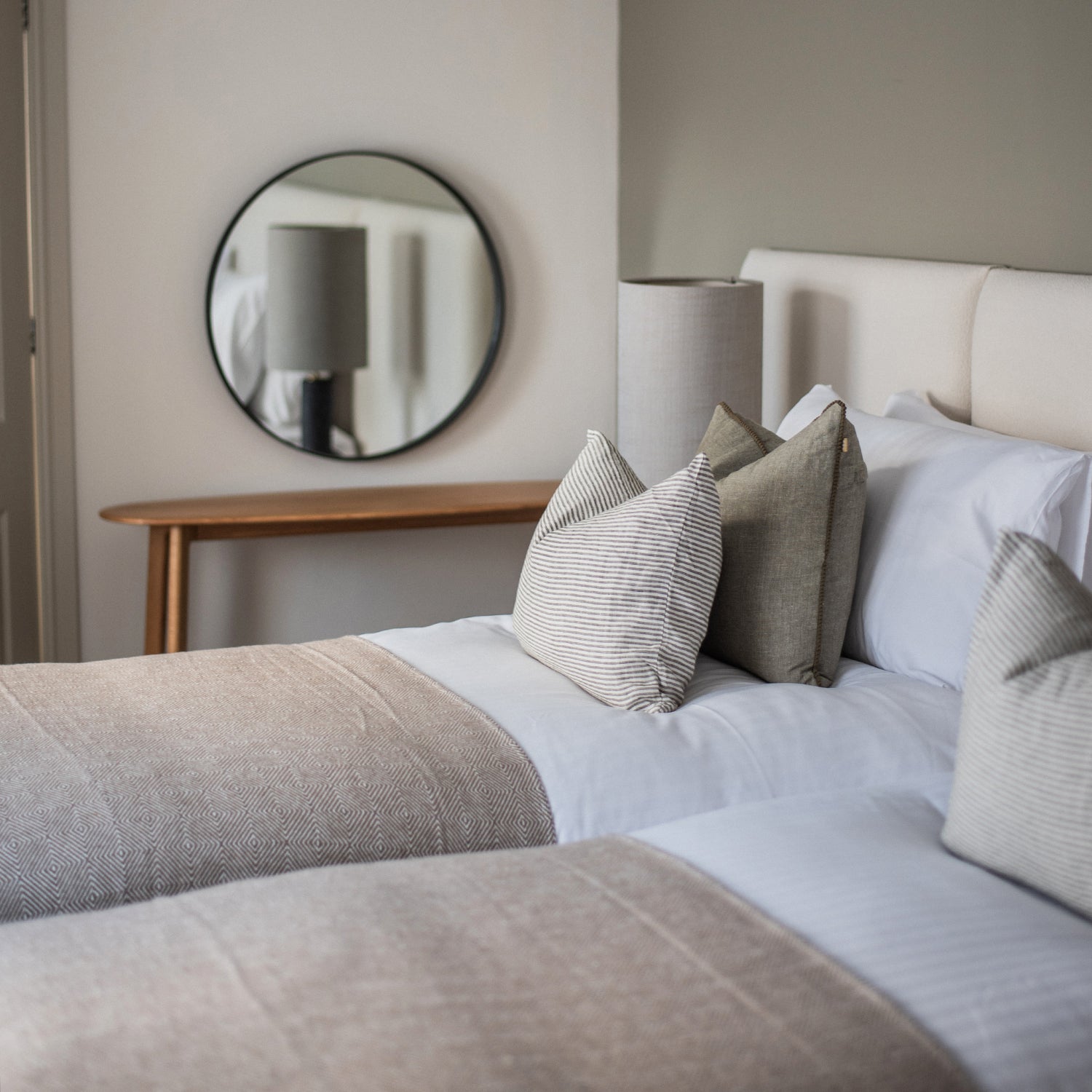 Elegant twin bedroom at Seaglass House featuring soft beige textiles, striped and linen cushions, a padded headboard, and a minimal wood console with a round wall mirror reflecting a floor lamp.