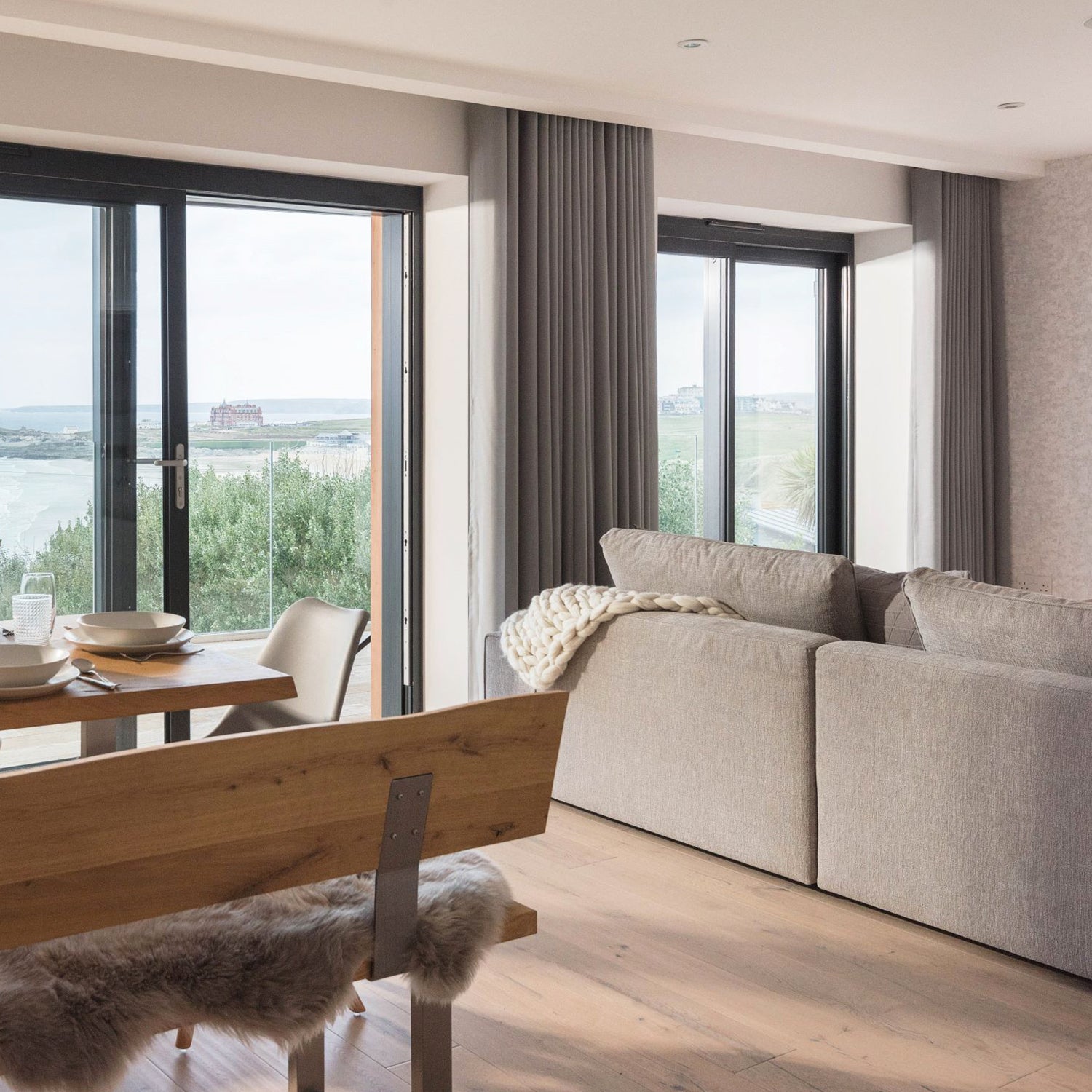 Contemporary living and dining area with full-length bespoke curtains on sliding glass doors, overlooking a coastal view — custom window dressings by luxury interior design experts.