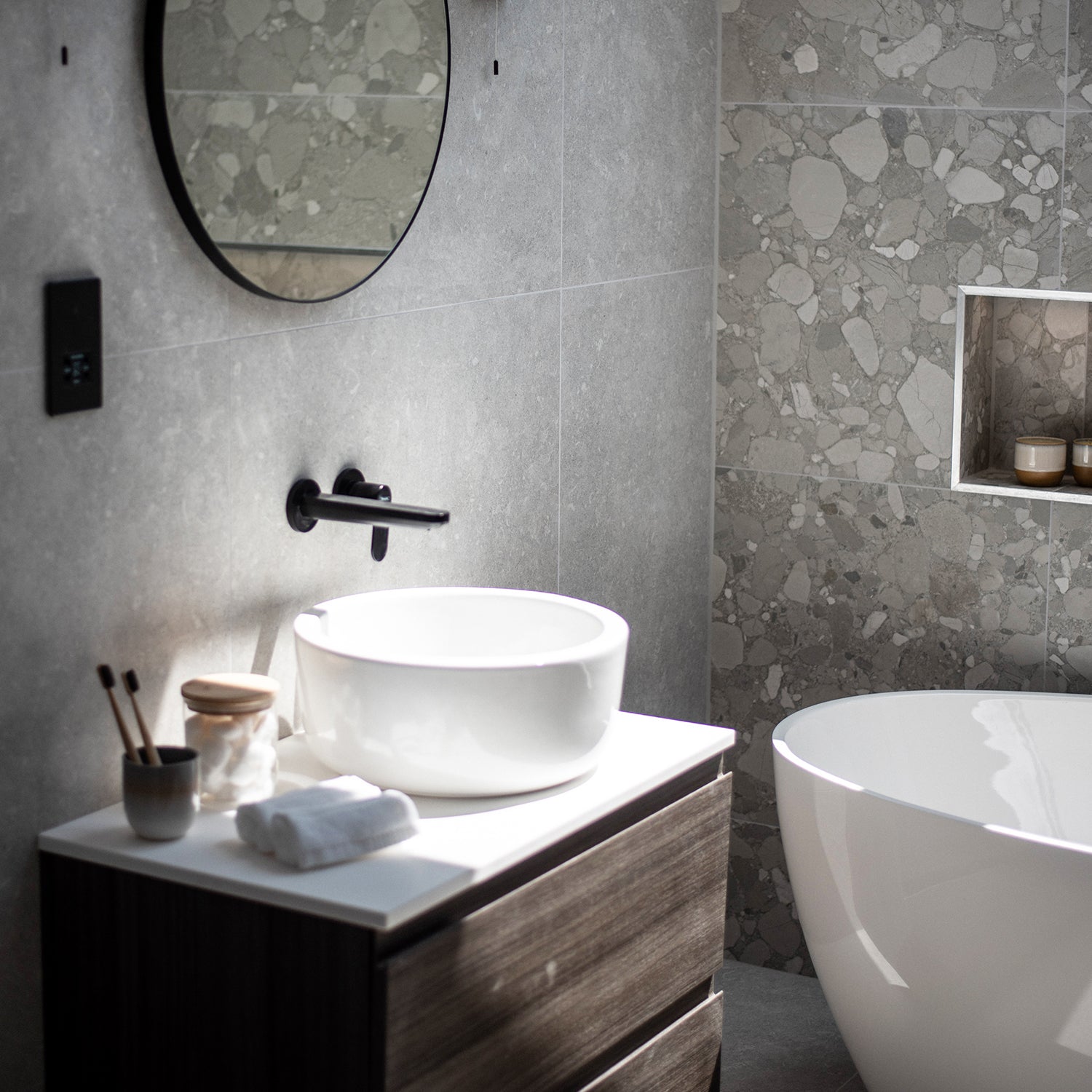 Modern rustic bathroom with concrete-effect walls, round mirror, freestanding white bathtub, and vessel sink on dark wood vanity – styled by Iroka Interiors.