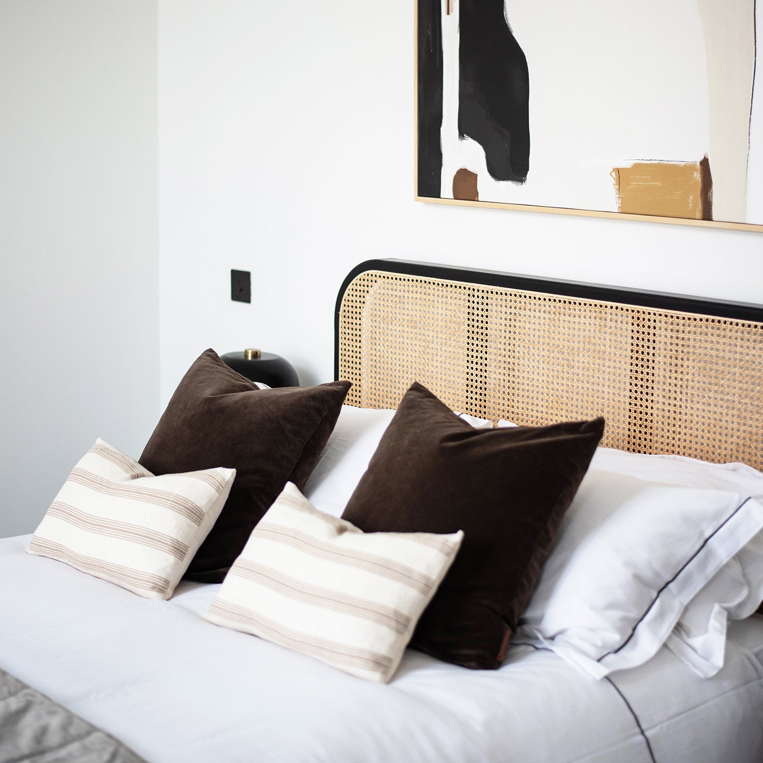 Contemporary bedroom with natural rattan headboard, layered cushions in earth tones, abstract wall art, and minimalist styling – interior design by Iroka in Playing Place, Truro.