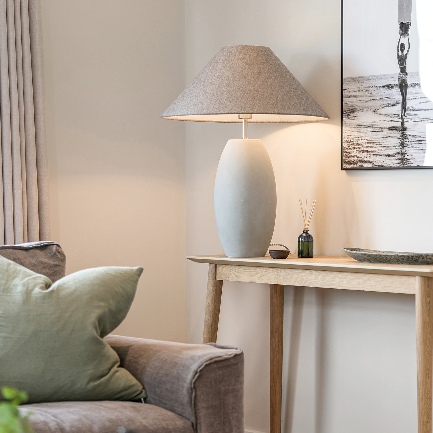 Modern living room detail with neutral ceramic table lamp, light oak console table, grey armchair with green cushion, and minimalist wall art – elegant contemporary interior styling with soft natural tones.