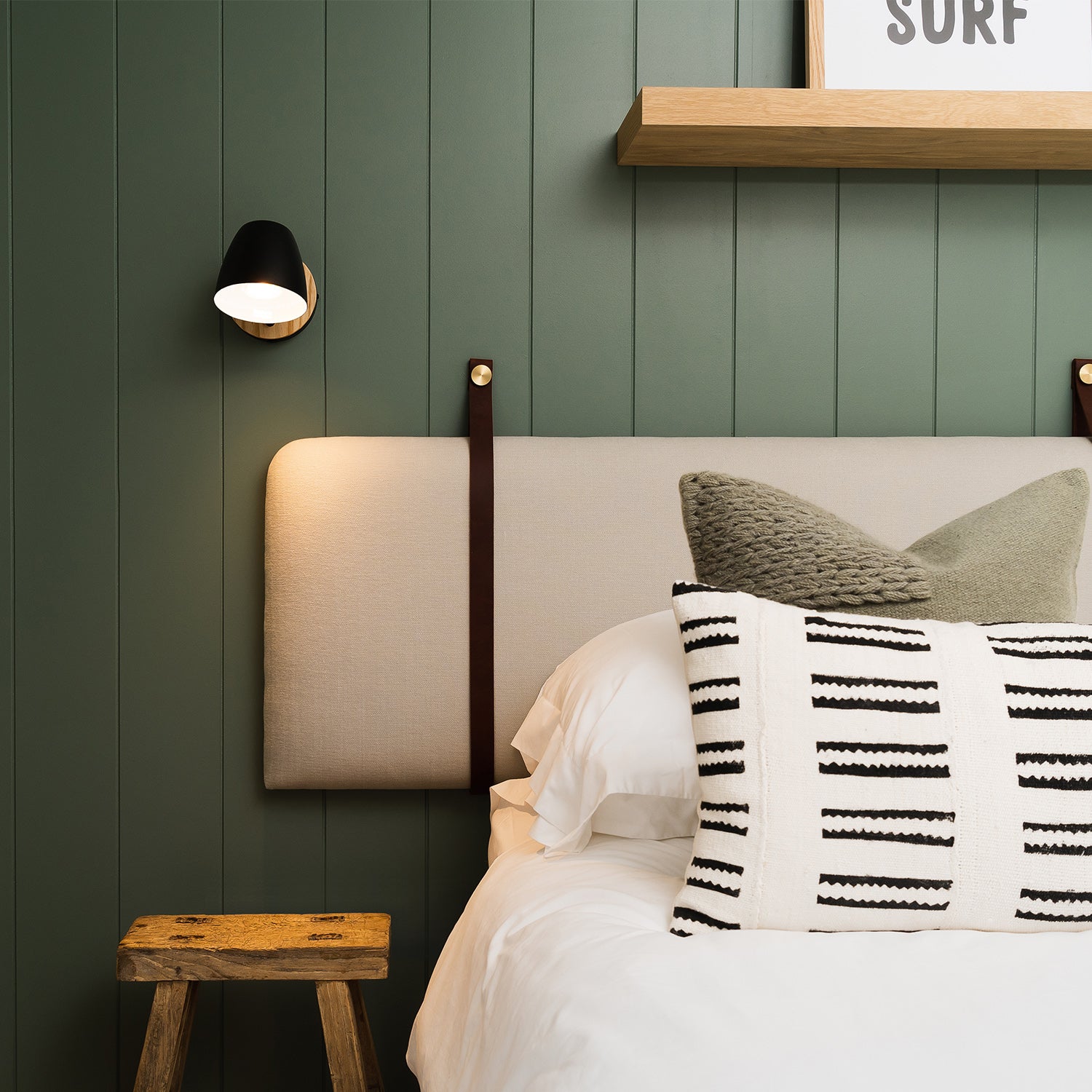 Stylish bedroom corner with green panelled wall, upholstered headboard, graphic cushions, black wall light, and oak stool – bespoke bedroom furniture and interior design by Iroka Interiors in Cornwall.
