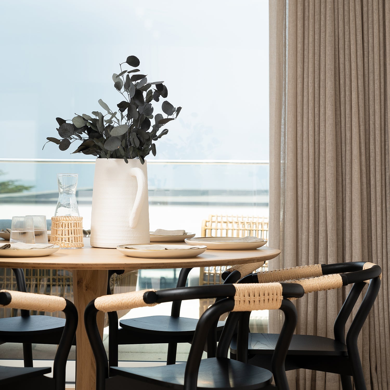 Scandinavian-inspired dining room featuring a light oak round table, modern black wishbone-style chairs with natural cord seats, and soft beige linen curtains — styled with minimalist tableware and a ceramic jug vase of eucalyptus.