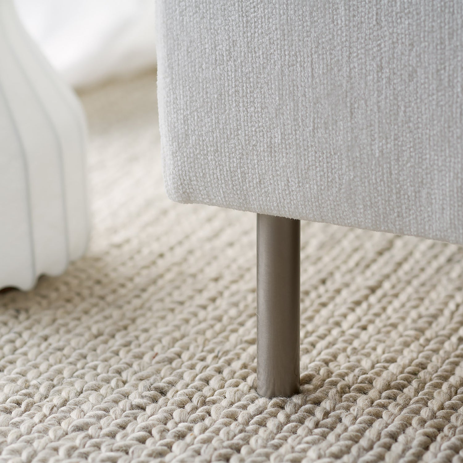 Close-up of slim metal sofa leg with off-white textured upholstery, resting on a chunky woven wool rug - highlighting Mastrella’s elevated and minimalist design details.