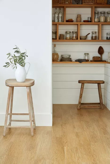 Warm oak-effect Amtico Form flooring in a rustic kitchen pantry with white shiplap walls, open shelving, and natural wood stools – showcasing a timeless and homely LVT finish.