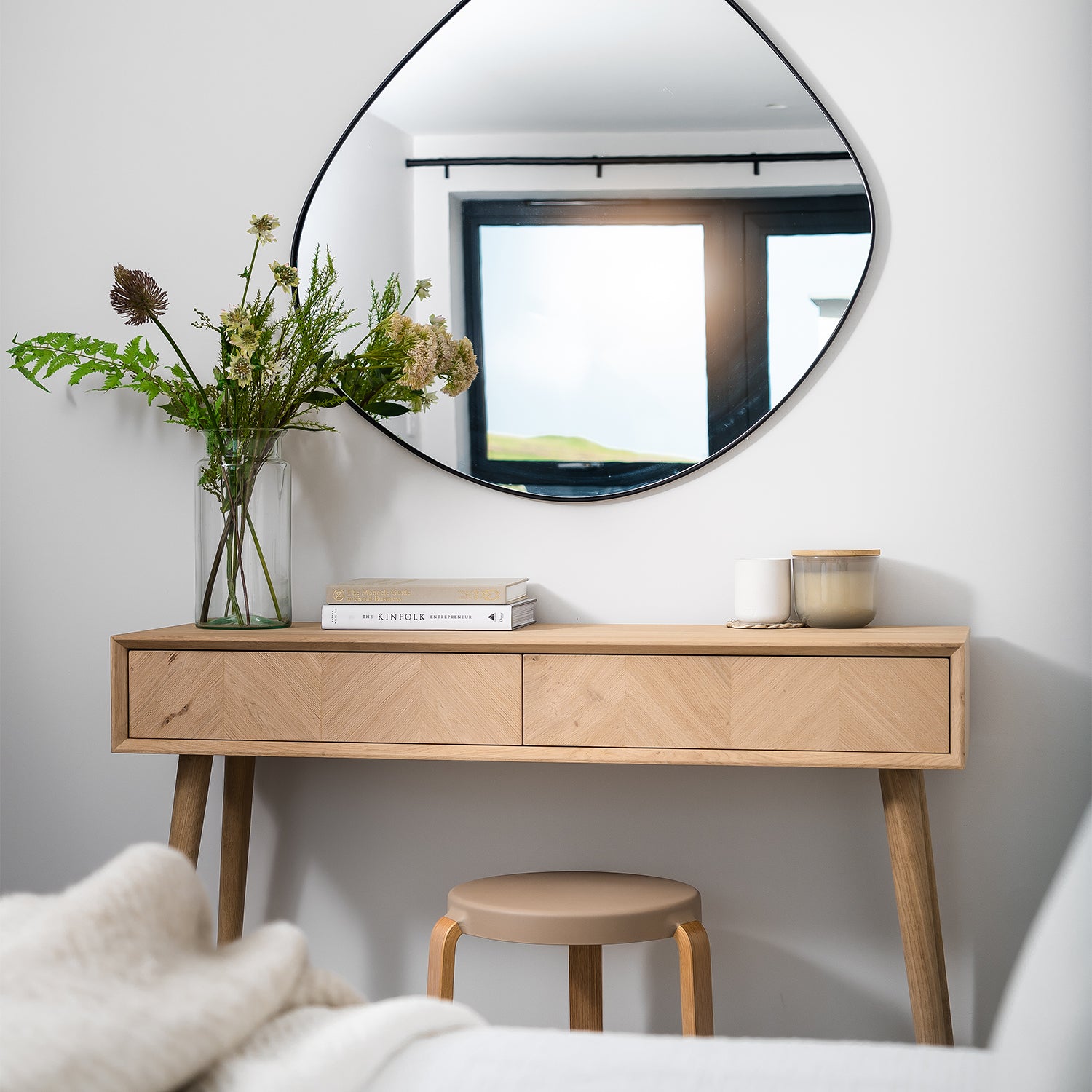 Scandinavian-style wooden dressing table with herringbone pattern, round wooden stool, asymmetrical mirror, vase of wildflowers, stacked books, and minimalist candles in a light-filled modern bedroom.