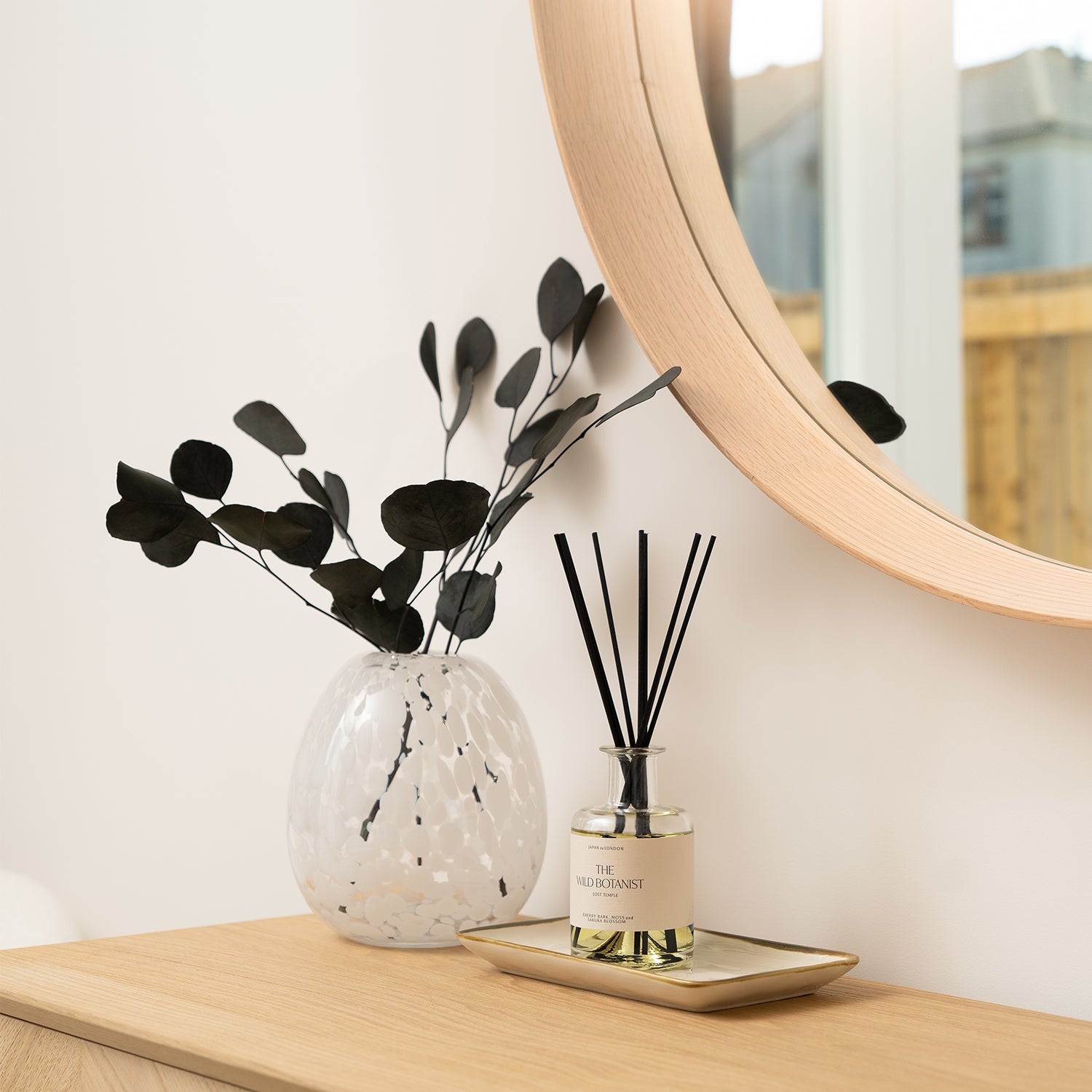 Modern home decor vignette with a round wooden mirror, white speckled vase holding black eucalyptus stems, and a luxury reed diffuser "The Wild Botanist" on a gold tray.