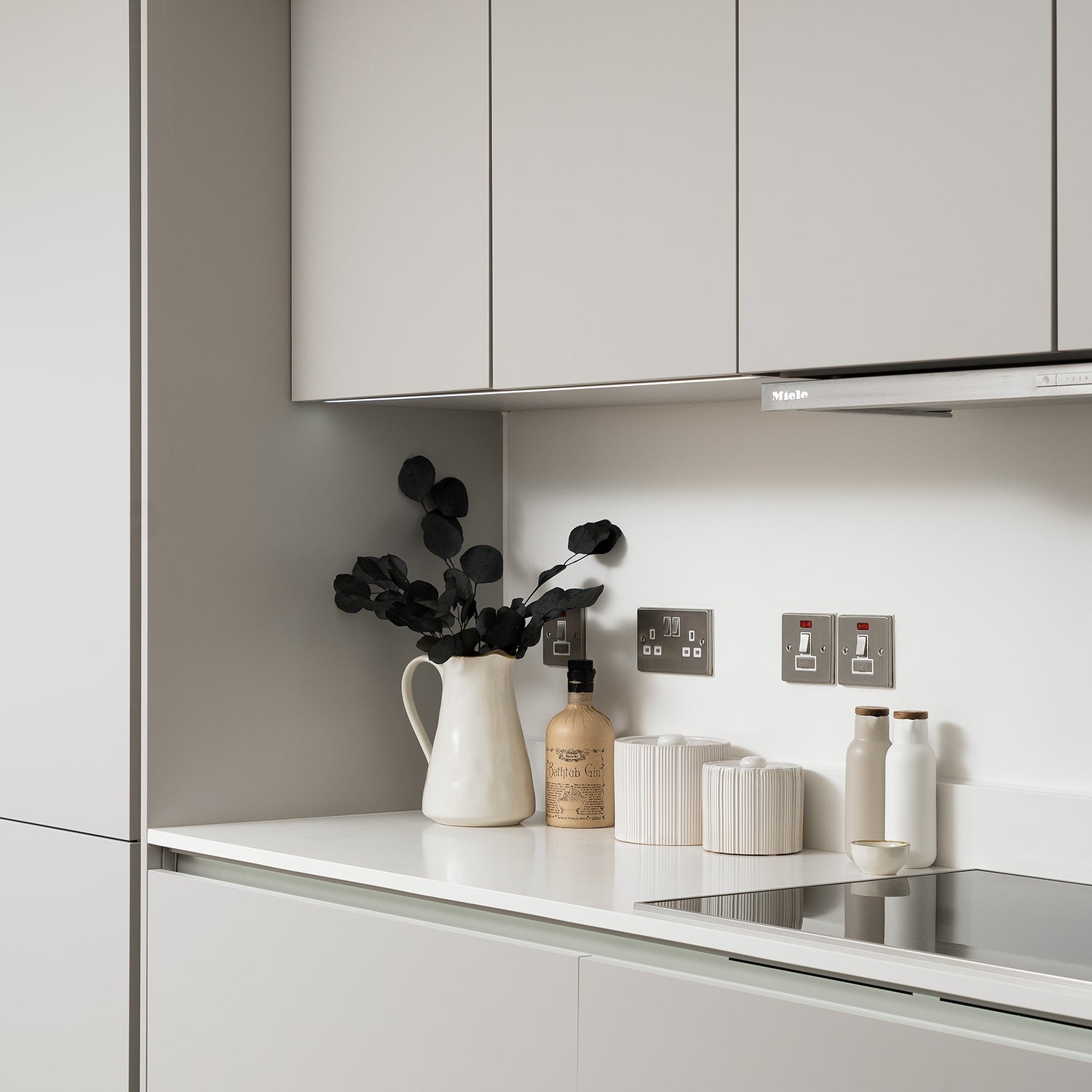 Modern minimalist kitchen countertop with neutral cabinetry, ceramic storage jars, decorative bottles, white pitcher with dark foliage, and sleek Miele extractor fan.